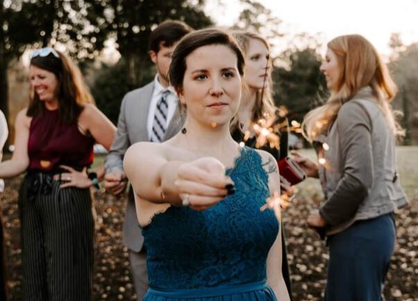 Photo of Ashton. She is at a wedding, holding a sparkler and looking off camera, smiling slightly.